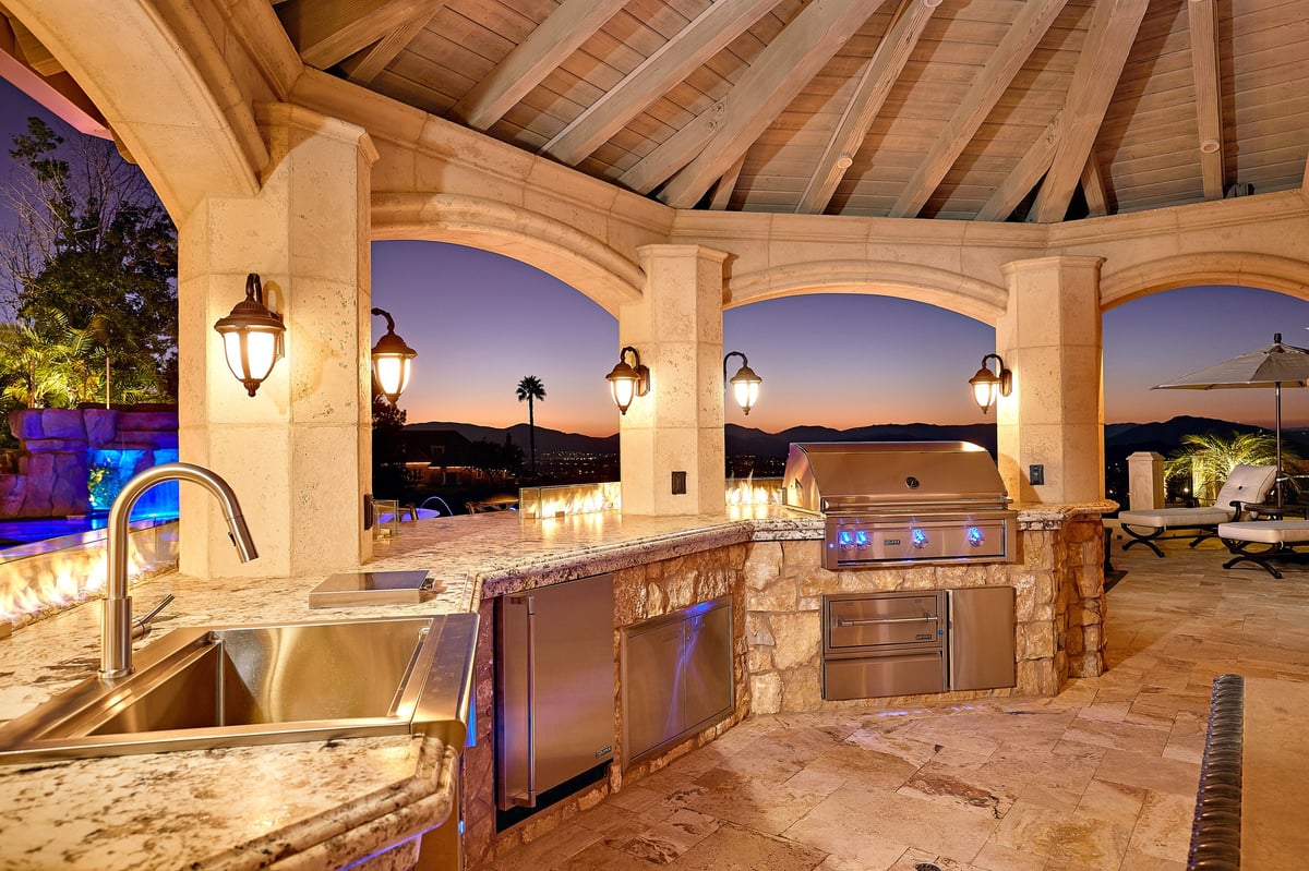 Luxurious outdoor kitchen at dusk with stone countertops, built-in grill, sink, and wooden pergola overhead, overlooking scenic desert landscape