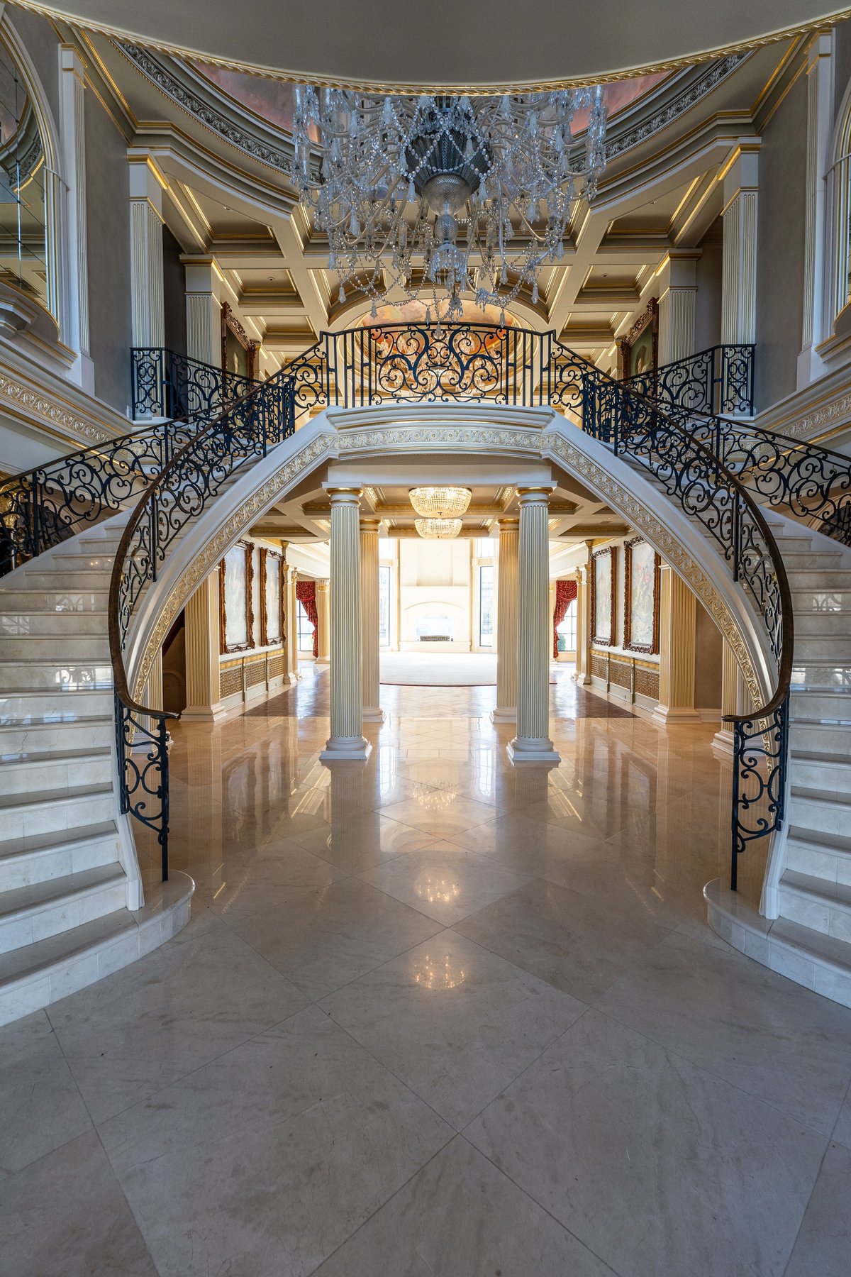 Grand elegant staircase with marble floors, ornate gold railings, white columns, and crystal chandelier in luxurious foyer