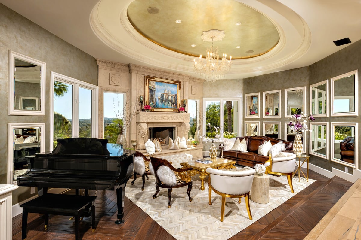 Luxurious living room with grand piano, elegant furniture, fireplace, chandelier, and large windows overlooking outdoors