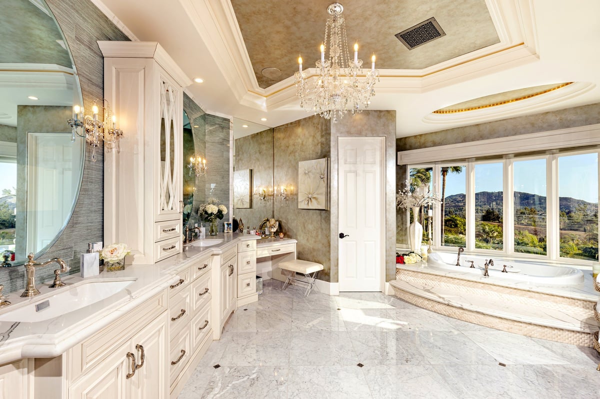 Luxurious master bathroom with white cabinetry, marble countertops, crystal chandelier, and soaking tub overlooking mountain views