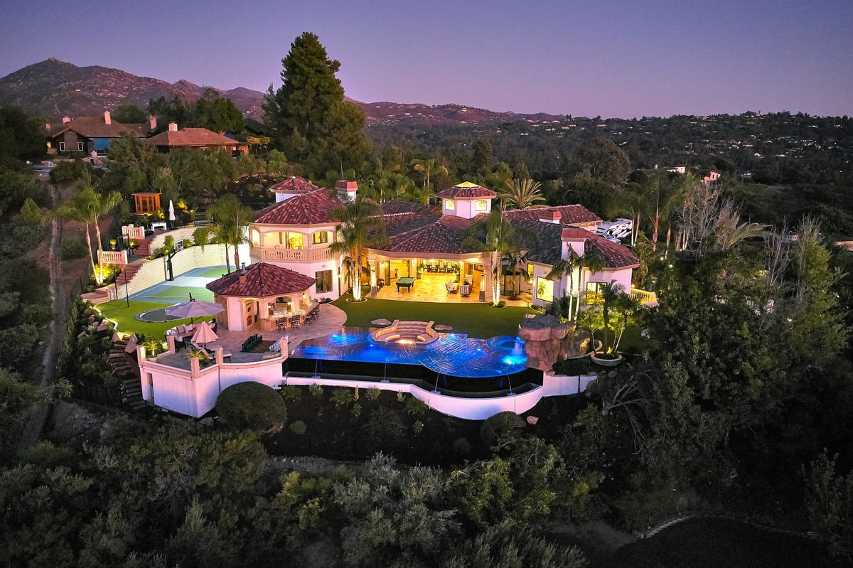 Aerial view of a luxury mansion with illuminated pink buildings, glowing swimming pool, and manicured grounds nestled in hillside vegetation at dusk