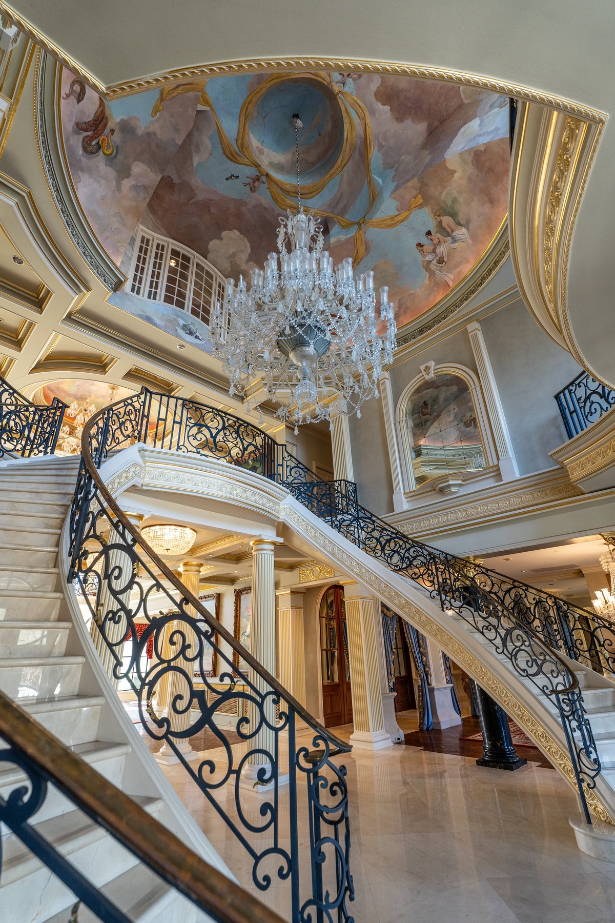 Grand staircase with ornate wrought iron railings beneath an elaborate frescoed ceiling dome and crystal chandelier