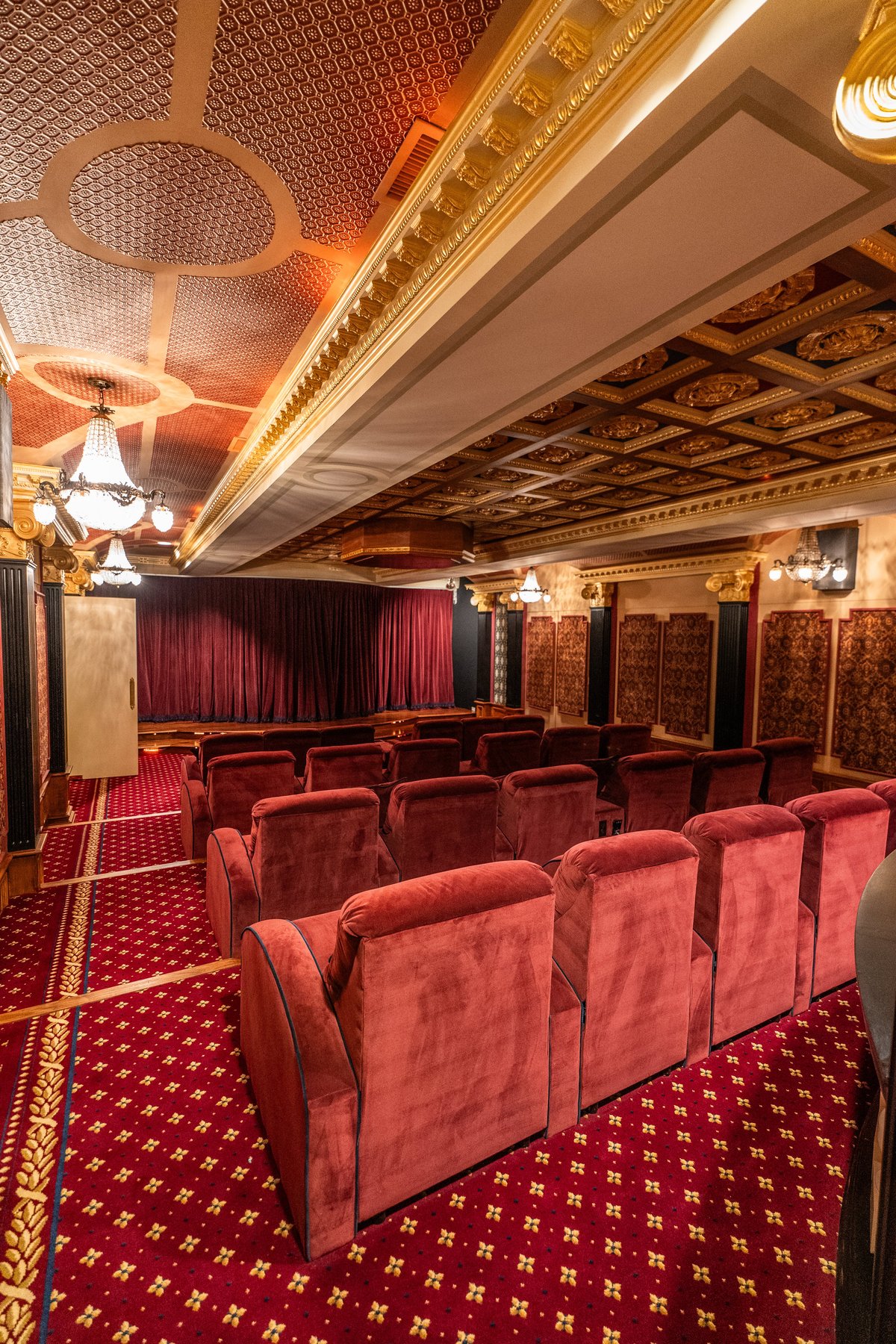 Elegant home theater with red velvet recliners, red curtained screen, ornate ceiling with gold trim, and red patterned carpet