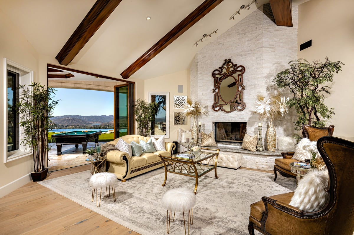 Luxurious living room with white exposed beams, fireplace, cream sofa, water view windows, potted plants, and elegant furnishings overlooking a lake