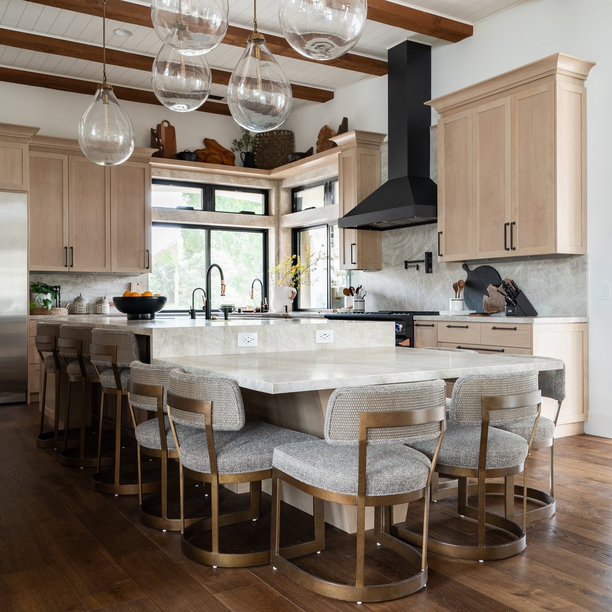 Modern kitchen with light wood cabinetry, large island with seating, marble countertops, glass pendant lights, and black range hood
