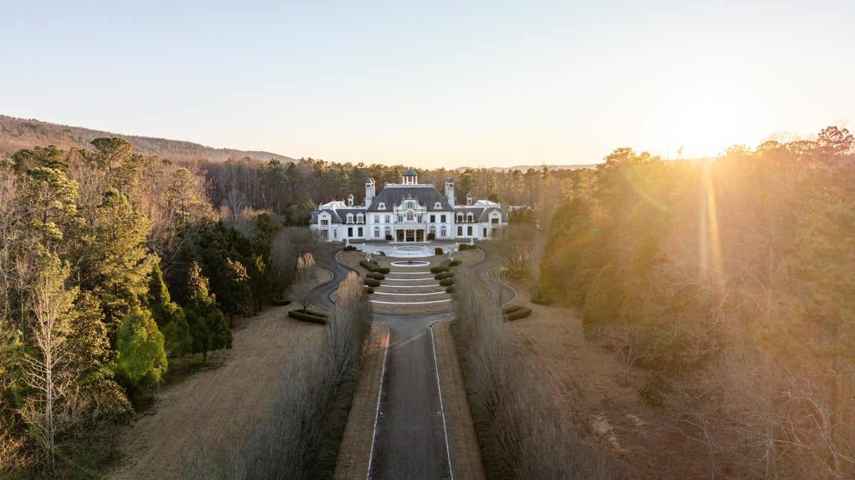 Aerial view of a grand white mansion with manicured gardens and tree-lined driveway at sunset