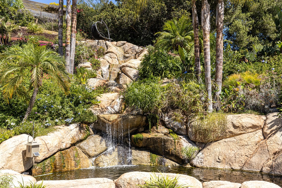 Natural waterfall flowing over moss-covered rocks surrounded by tall palms and lush green vegetation in a tropical garden setting